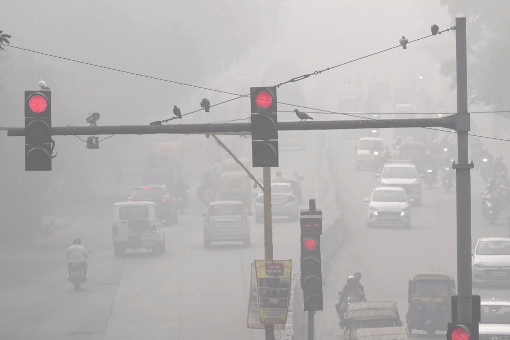 Dense winter fog over Delhi‑NCR road with vehicles using headlights due to low visibility.