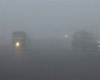 Vehicles and commuters moving through dense cold fog across North Indian plains under winter weather conditions.